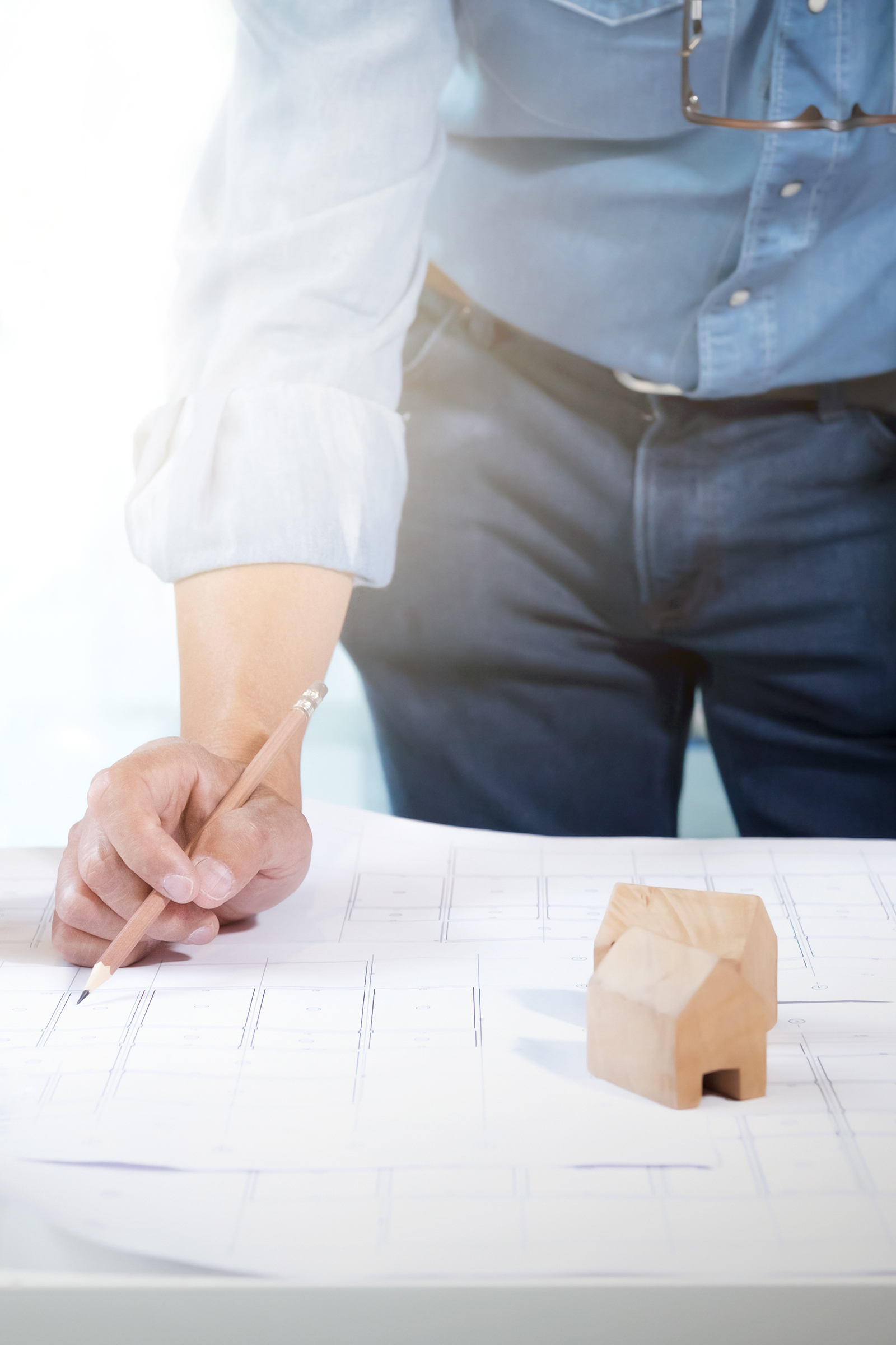 Foto van een architect met een potlood in zijn hand. Een bouwplan is duidelijk zichtbaar op tafel. Ten slotte staan er twee houten miniatuur huisjes op dat bouwplan.
